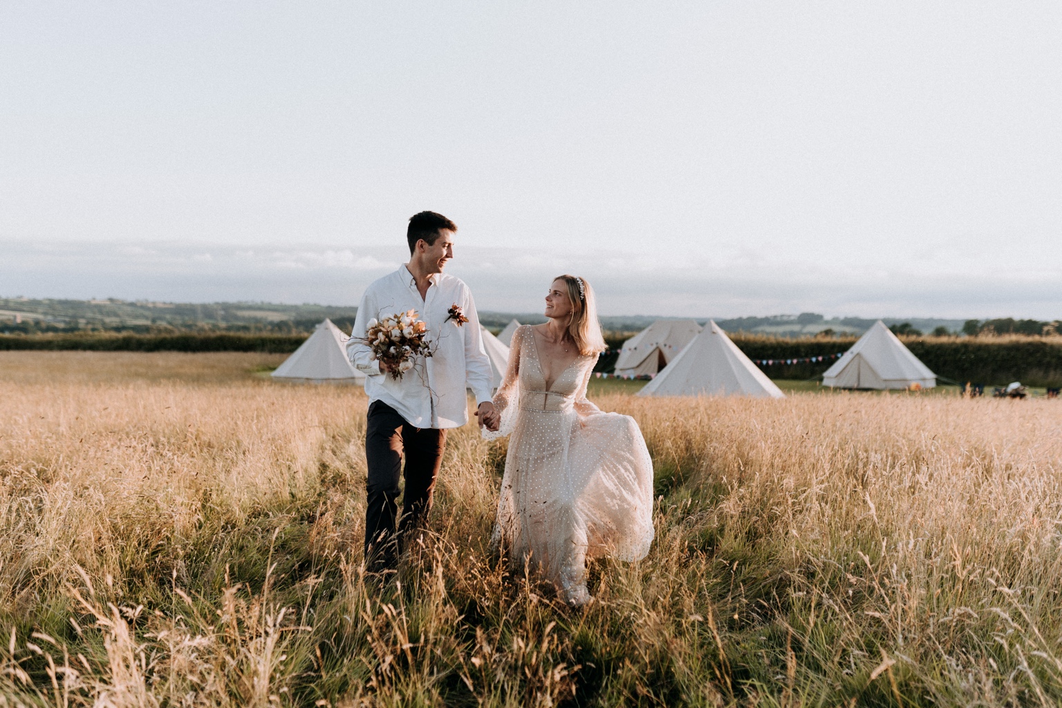 Bride and groom holding hands as they walk through tall grass, bell tents behind