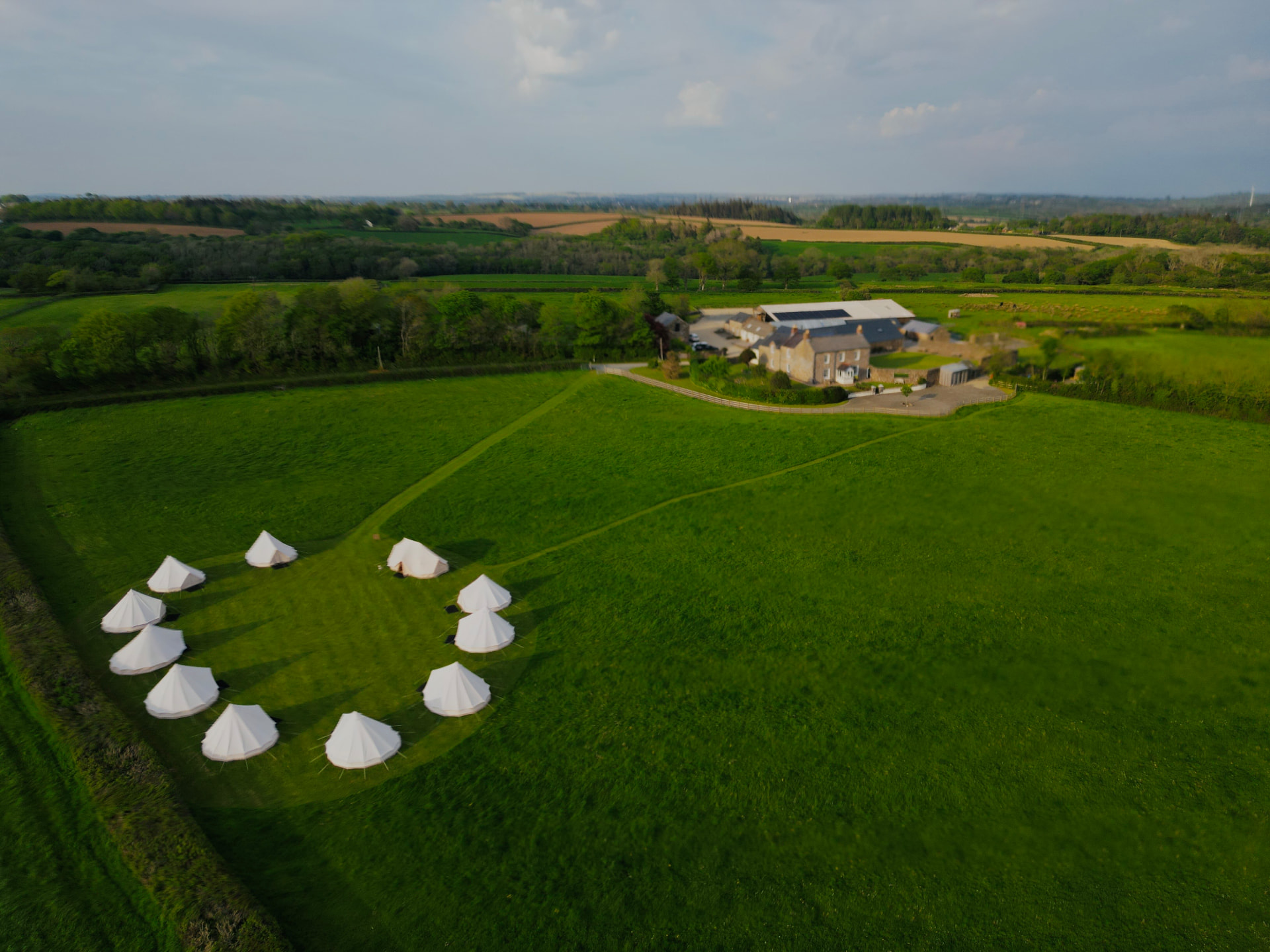 Aerial view of eleven white bell tents arranged in a wide circle on a green field beside a stone farmstead