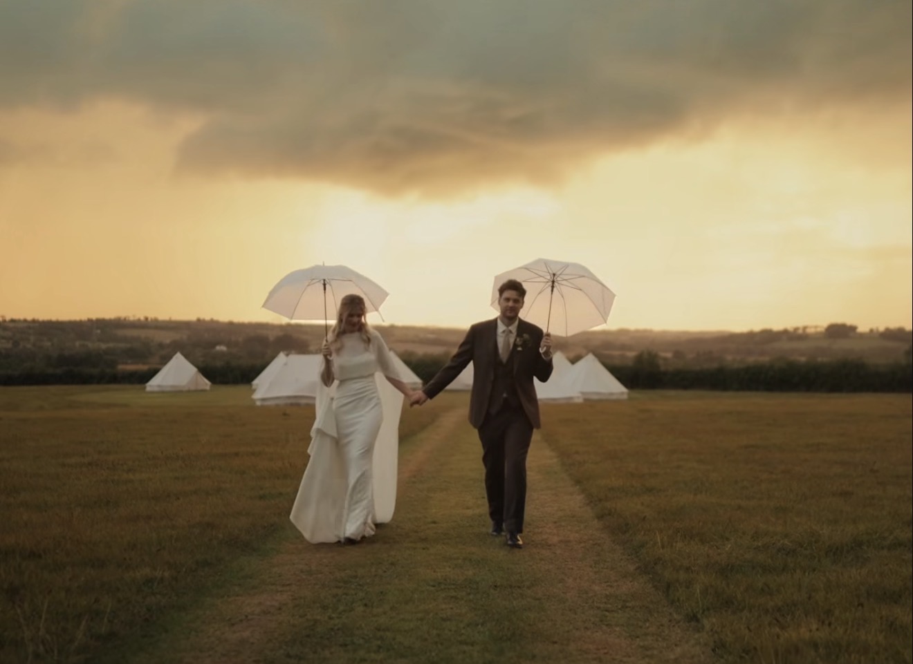 Bride and groom walking with umbrellas at sunset, bell tents in the distance