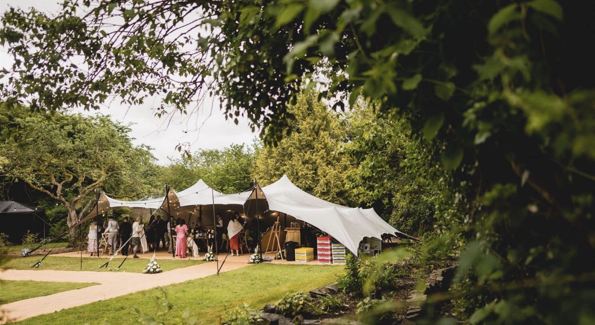 Open-sided stretch tent framed by tree branches, with guests gathered on the lawn