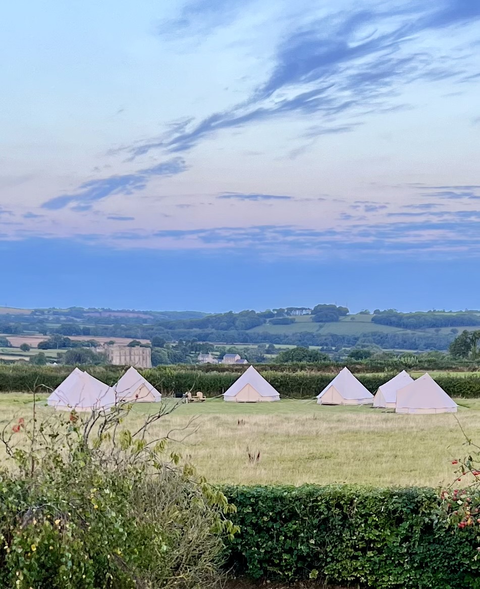 Row of bell tents in a field overlooking hills and historic buildings at dusk