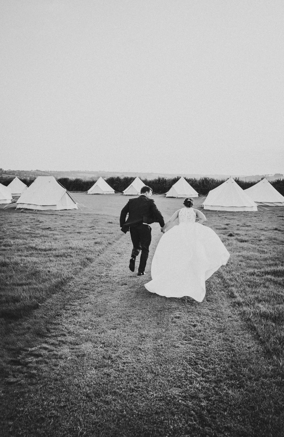 Black-and-white photograph of a bride and groom running hand-in-hand towards bell tents