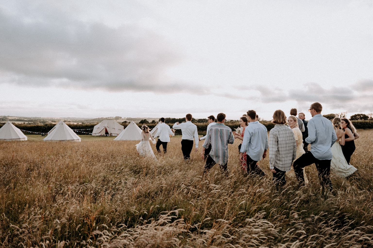 Wedding guests and a bride crossing a tall grass field towards white bell tents