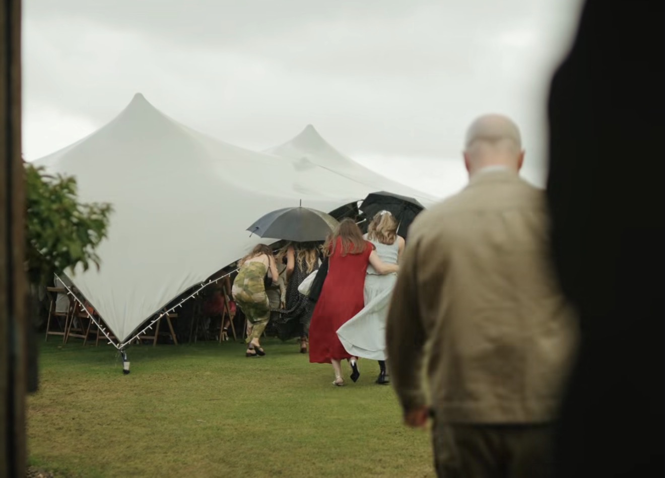Guests with umbrellas crossing a lawn towards a white marquee on a rainy day